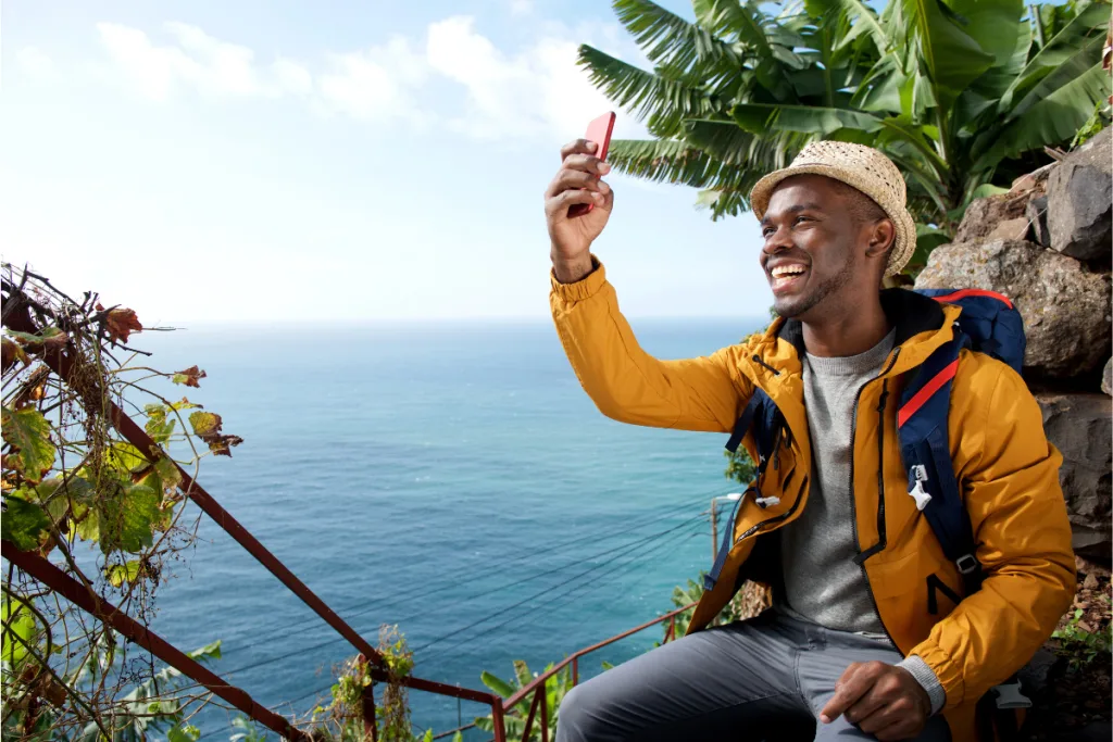 Benefits of Solo Travel, A young man in a yellow jacket and hat taking a selfie with a smartphone near the ocean.