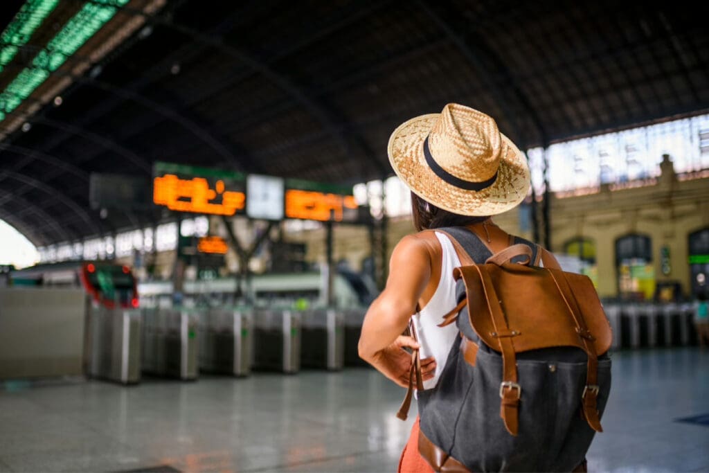 Travel on a Whim, A solo traveler wearing a straw hat and carrying a backpack stands at an airport gate looking at departure information.