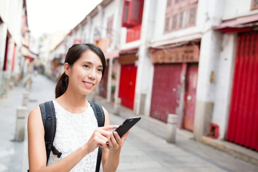 Best Travel Application,Young woman with a backpack using a smartphone while standing in a street with red storefronts.