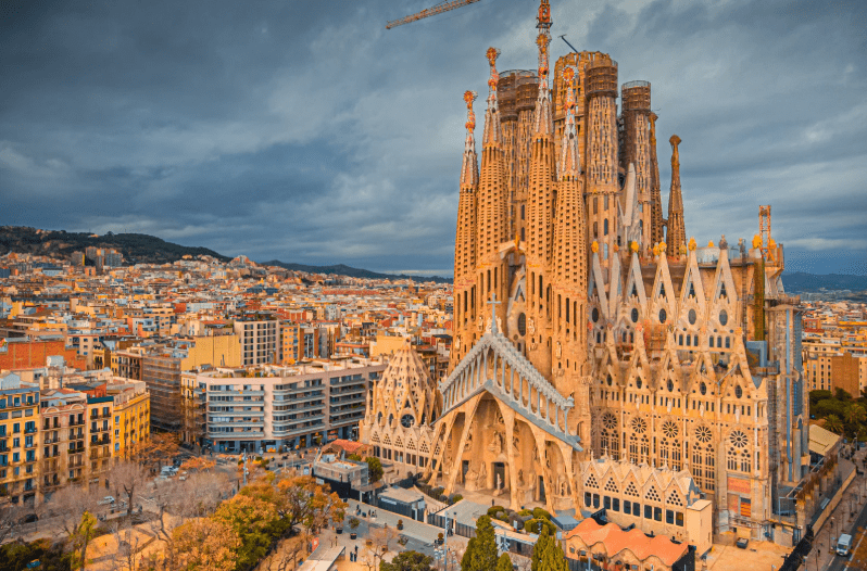 Aerial view of the Sagrada Familia basilica, one of the UNESCO World Heritage Sites in Spain, with its ornate towers and facades under construction, surrounded by Barcelona’s city buildings beneath a cloudy sky.