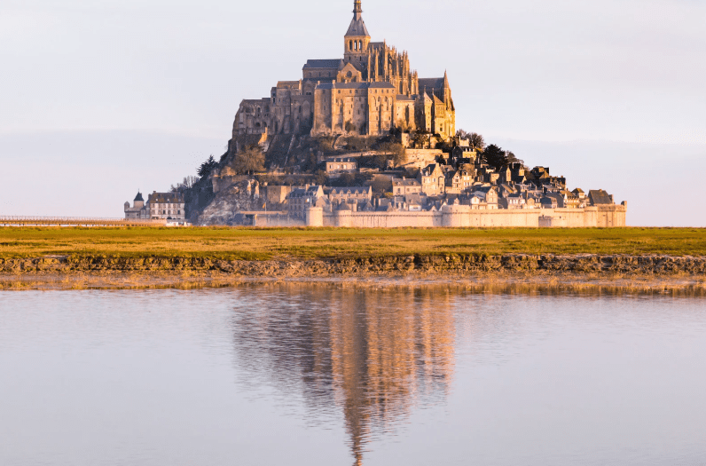 A historic stone abbey and village stand atop a rocky island, reflected in calm water below, with a grassy area in the foreground and a clear sky in the background.