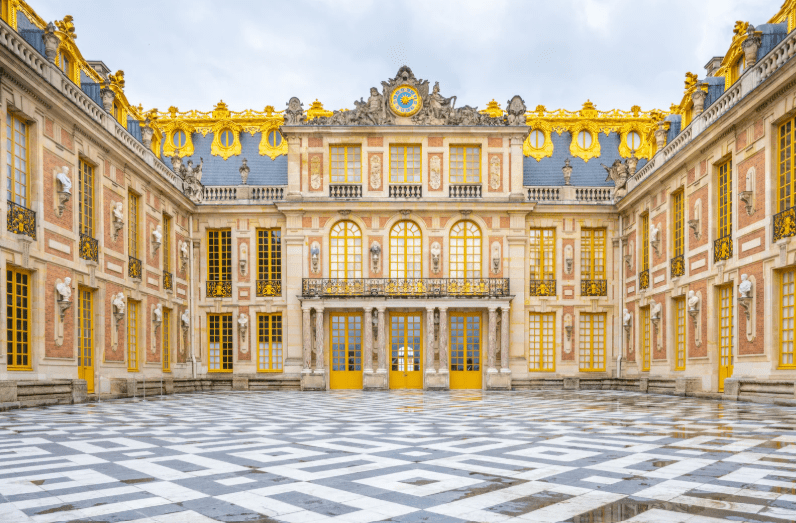 The photo shows the elegant marble courtyard of the Palace of Versailles, with gold-accented windows, ornate details, and a dramatic black-and-white checkerboard floor under a cloudy sky.