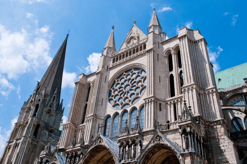 The image shows the ornate facade of a Gothic cathedral with a tall spire, pointed arches, and a large circular rose window under a bright blue sky with scattered clouds.