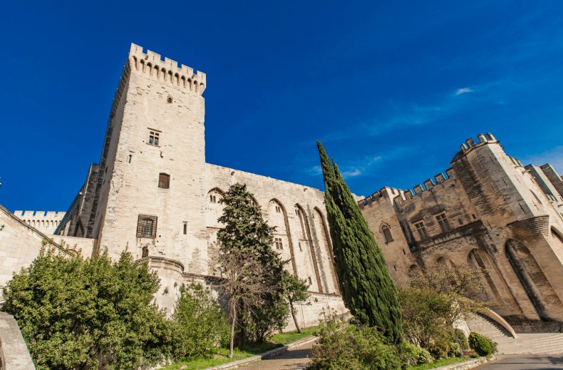 A historic stone castle with tall towers and crenellated walls, surrounded by green trees and shrubs, stands under a clear blue sky.