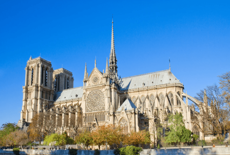 Notre-Dame Cathedral in Paris, France, with its iconic Gothic architecture, rose windows, and twin towers, stands under a clear blue sky, surrounded by trees with autumn foliage.