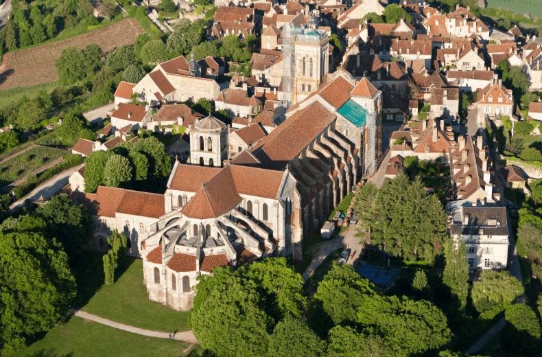 Aerial view of a historic European town—reminiscent of UNESCO World Heritage Sites in France—with a large stone church featuring red-tiled roofs, nestled among trees, smaller houses, and winding streets.