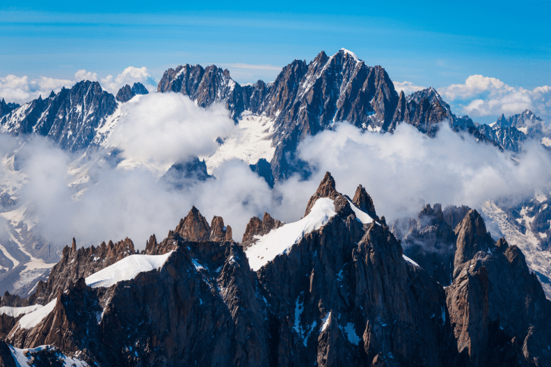 Jagged, snow-capped mountain peaks—part of the UNESCO World Heritage Sites in France—rise above the clouds under a bright blue sky, with rugged rock formations and patches of snow visible in the foreground.