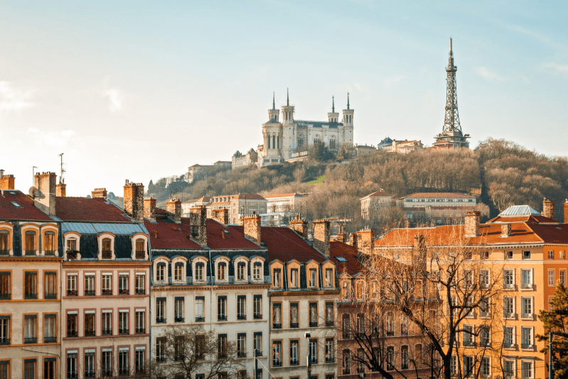 A panoramic view of Lyon, France, a gem among UNESCO World Heritage Sites in France, with colorful historic buildings in the foreground and the Basilica of Notre-Dame de Fourvière and metallic tower atop a hill under a clear sky.