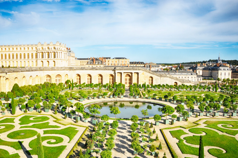 The photo shows the ornate gardens of the Palace of Versailles, one of the UNESCO World Heritage Sites in France, featuring manicured hedges, symmetrical patterns, fountains, and classical architecture under a bright blue sky.