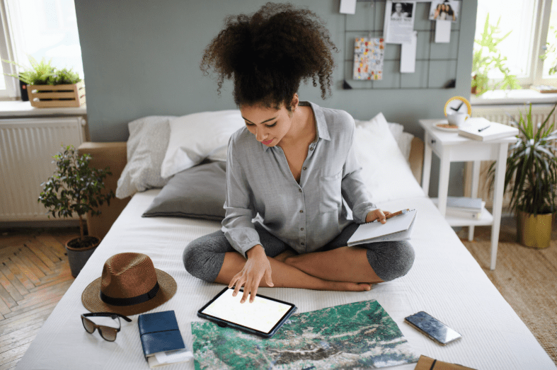 A woman sits cross-legged on a bed using a tablet, surrounded by travel essentials like a map, passport, hat, sunglasses, and a notebook, appearing to plan a trip to explore UNESCO World Heritage Sites in France in her bright, cozy room.