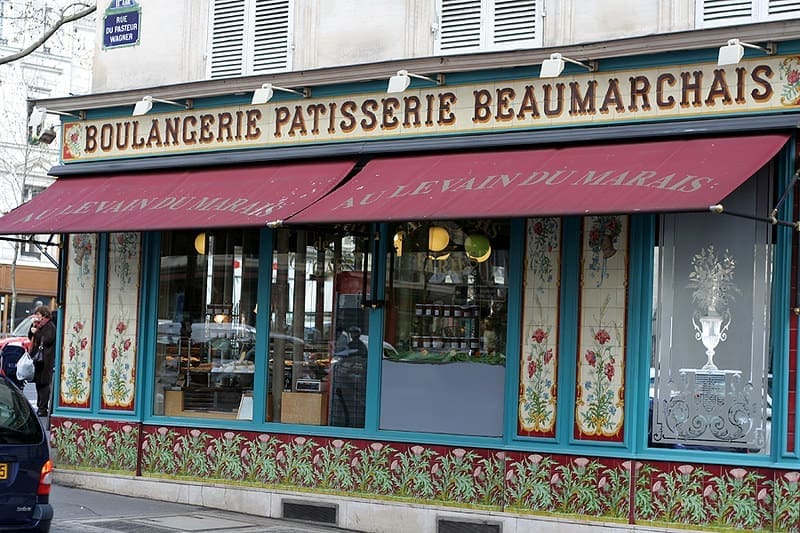 A French bakery and pastry shop, Boulangerie Patisserie Beaumarchais, with red and green decorative awnings and large windows displaying common food in France like fresh bread and pastries. People are passing by on the sidewalk outside.