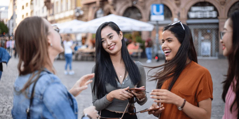 Four young women stand outside in a city square, smiling and laughing together while holding their phones. Cafés and historic buildings are visible in the background.