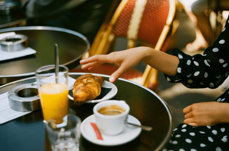 A person in a black and white polka dot dress reaches for a croissant—a common food in France—on a plate at an outdoor café table, with a cup of coffee, a glass of orange juice, and an ashtray nearby.