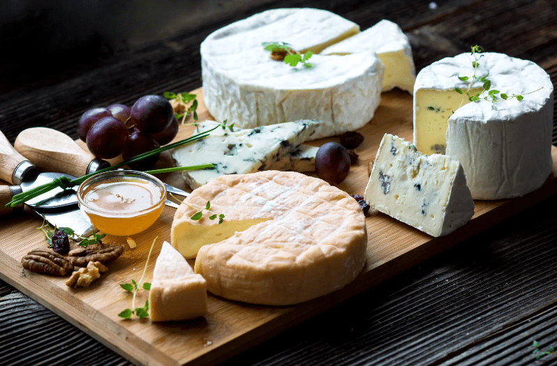 A wooden board with assorted cheeses—a common food in France—grapes, pecans, walnuts, a small glass bowl of honey, a cheese knife, and fresh herb garnishes, set on a rustic wooden surface.