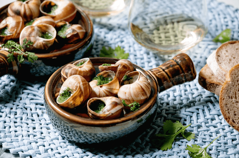 A ceramic dish of cooked escargot, a common food in France, is garnished with herbs and sits on a woven placemat, accompanied by slices of bread and glasses of white wine in the background.