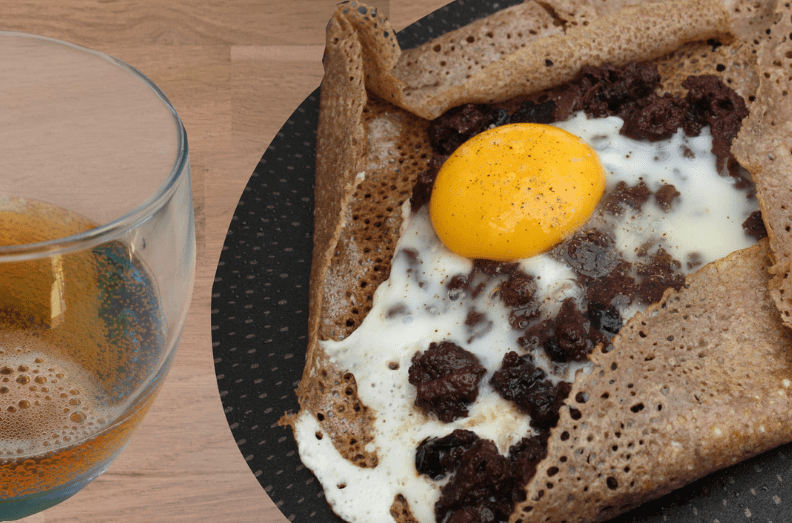 A common food in France, this buckwheat crepe is topped with a sunny-side-up egg and crumbled sausage on a black plate, served beside a glass of cider on a wooden table.
