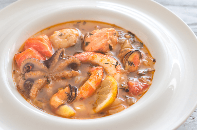 A white bowl filled with seafood soup, a common food in France, featuring shrimp, octopus, mussels, salmon, lemon slices, and herbs in a clear broth.
