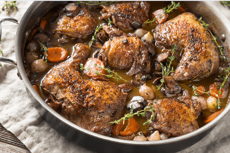 A large pan filled with braised chicken thighs, carrots, pearl onions, mushrooms, and fresh herbs in a rich brown sauce—this common food in France is cooked and garnished with sprigs of fresh thyme.