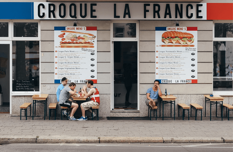 Four people sit at two outdoor tables in front of a restaurant called Croque La France. Large menu boards with common food in France, including sandwich options, are displayed on the wall behind them. One person sits alone, while three others sit together.