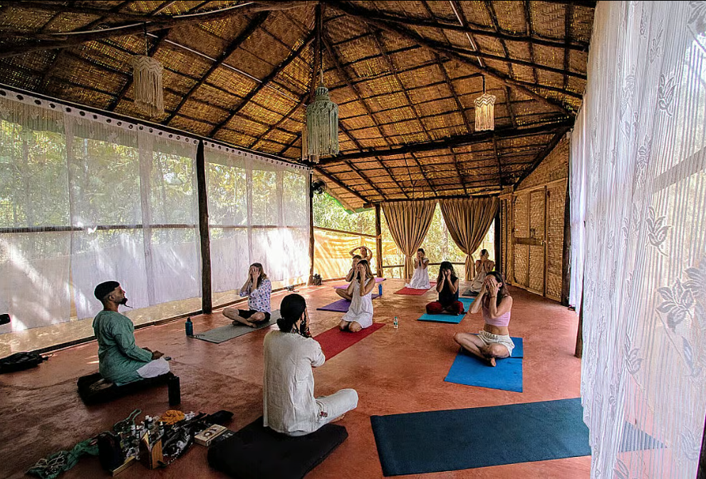 A group of people sit cross-legged on yoga mats inside an open, bamboo-roofed pavilion with sheer curtains, participating in a guided meditation or yoga session—reminiscent of meditation retreats in India—led by two instructors.