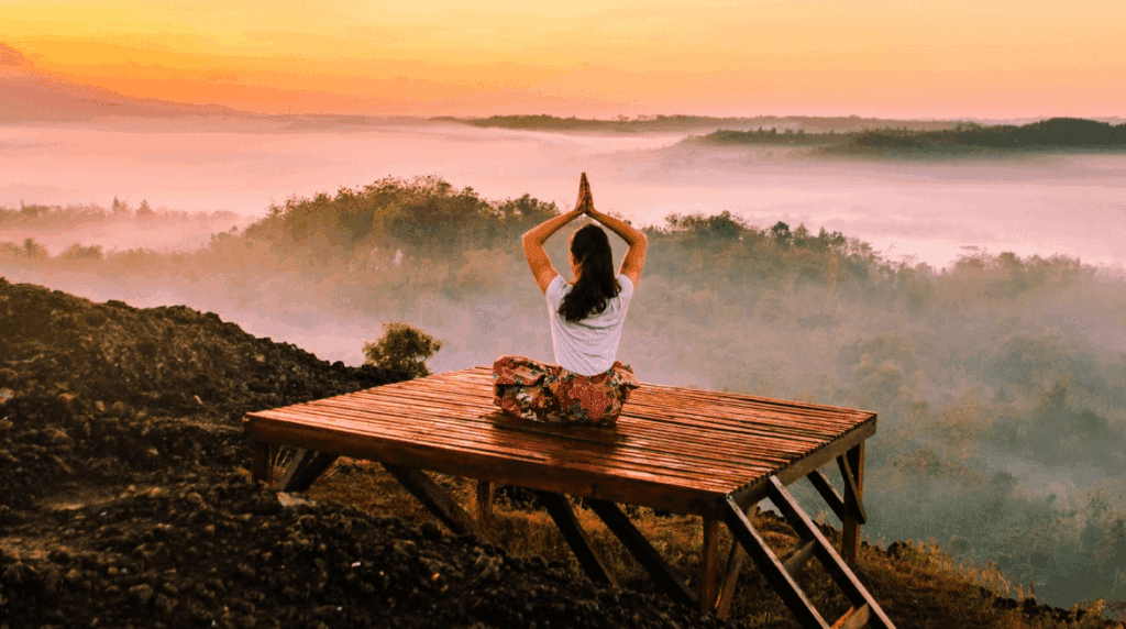 Meditation retreats in India, A person sits cross-legged on a wooden platform, facing a misty sunrise landscape with hands raised in a yoga pose—capturing the peaceful, serene atmosphere often found at meditation retreats in India.