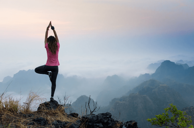 A person stands on a rocky peak, balancing in a yoga tree pose at sunrise or sunset, overlooking misty mountains and a valley below—a scene reminiscent of Yoga Retreats in Costa Rica.