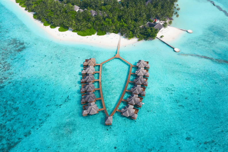 Aerial view of overwater bungalows arranged in a fish shape above clear turquoise water, with a white sandy beach, lush green palm trees, and more huts along the shore—truly one of the prettiest beaches in the world.