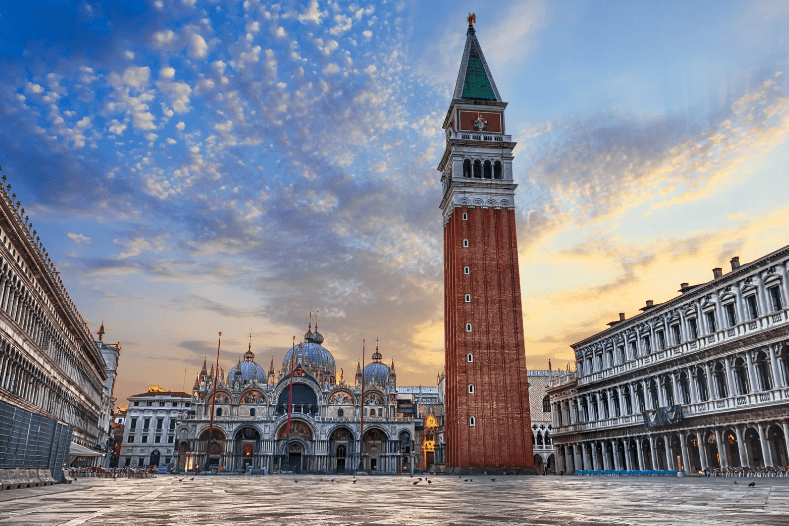 St. Mark’s Square in Venice, Italy, at sunrise, is a highlight among UNESCO World Heritage Sites in Italy, featuring St. Mark’s Basilica with its domes and the grand Campanile bell tower, surrounded by historic buildings under a partly cloudy sky.