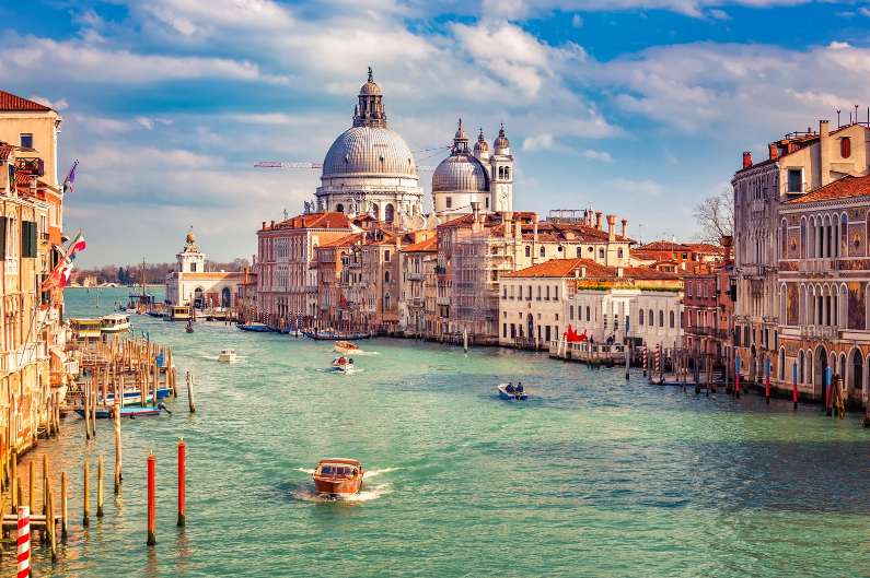 UNESCO World Heritage Sites in Italy, A scenic view of Venice’s Grand Canal, part of the UNESCO World Heritage Sites in Italy, with boats on turquoise water, historic buildings on both sides, and the domed Santa Maria della Salute basilica under a partly cloudy sky.