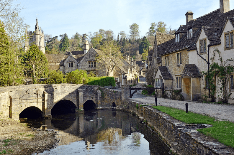 A stone bridge crosses a narrow stream in a quaint village—one of the hidden gems in England—with old stone houses, a church with a tall spire, and trees set against a clear sky.