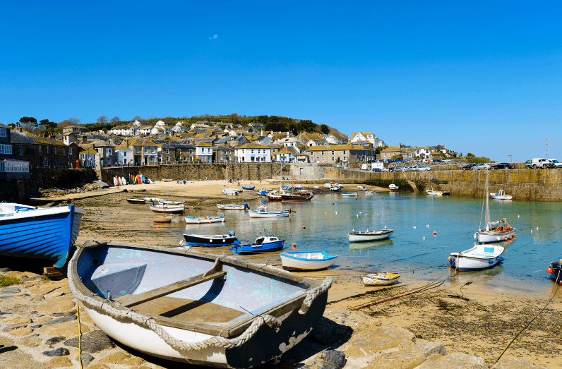 Small boats rest in a sunny harbor with calm water, surrounded by a stone wall. Houses dot the hillside in the background under a clear blue sky—one of the hidden gems in England waiting to be discovered.