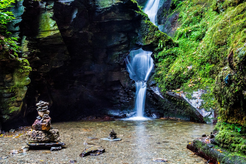 A small waterfall cascades down mossy rocks into a clear pool, surrounded by lush greenery. Rock cairns are stacked at the water’s edge—one of those hidden gems in England waiting to be discovered.