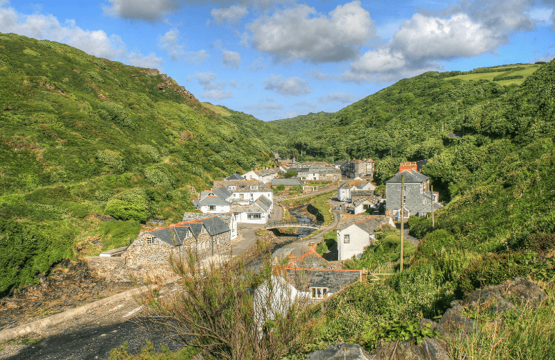 Hidden Gems in England, A picturesque village, one of the hidden gems in England, nestles in a lush green valley with stone and white houses, a small river, and hills covered in dense vegetation beneath a partly cloudy blue sky.