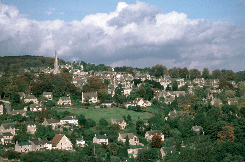 A picturesque village with stone houses, trees, and gardens spreads across rolling hills—a true hidden gem in England. A tall church steeple rises in the background under a partly cloudy sky.