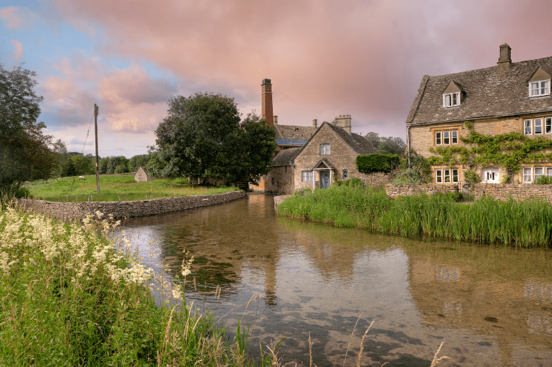 A serene village scene, one of the hidden gems in England, with stone houses beside a clear, shallow stream, lush greenery, and a partly cloudy pinkish sky at sunset. Wildflowers grow along the stream’s edge and a stone wall curves along the water.