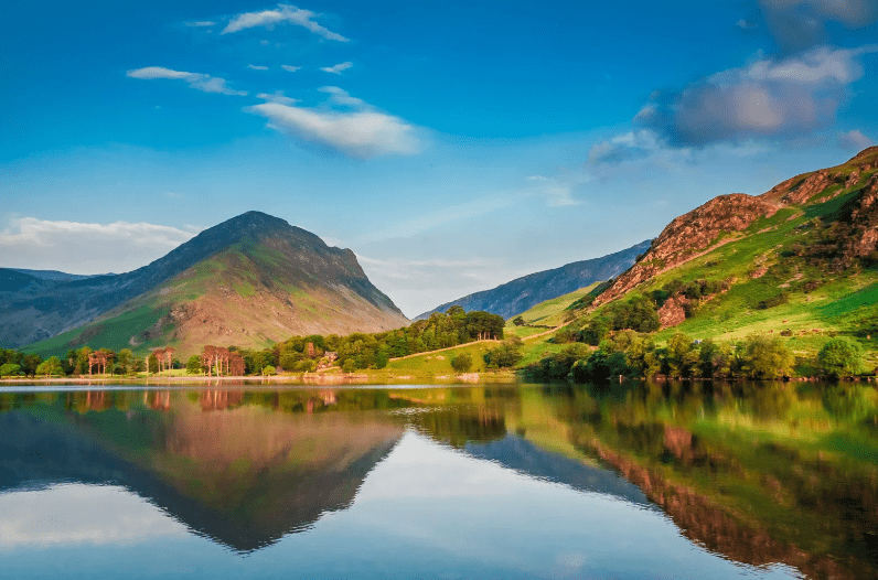 A calm lake reflects green hills and a tall mountain under a blue sky with scattered clouds, creating a serene and peaceful landscape—one of the hidden gems in England waiting to be discovered.