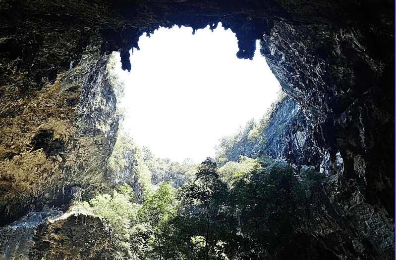 A large cave opening reveals sunlight, rocky walls, and lush green trees outside. The dramatic contrast of bright sky and shadowy cave showcases one of the hidden gems in England’s remarkable natural landscape.
