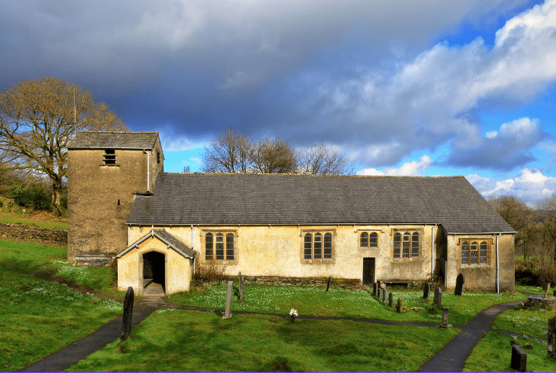 An old stone church with a bell tower stands in a grassy cemetery, surrounded by gravestones and trees under a partly cloudy sky. Sunlight highlights the weathered walls and slate roof of one of the hidden gems in England.