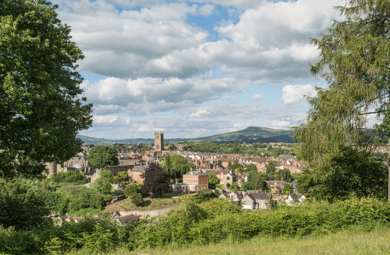 A scenic view of a small town, one of the hidden gems in England, with historic buildings and a prominent church tower, surrounded by lush greenery and hills under a partly cloudy sky.