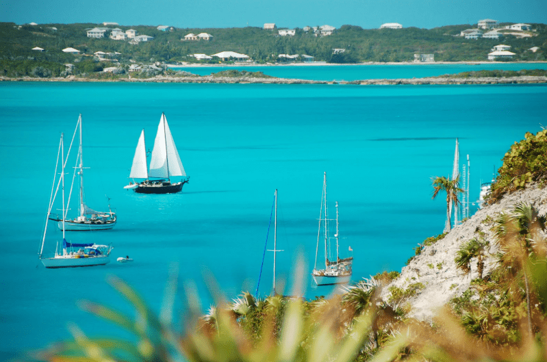 Sailboats with white sails float on vibrant turquoise water near a tropical shoreline, with palm trees and island houses—capturing the essence of the prettiest beaches in the world under a clear blue sky.