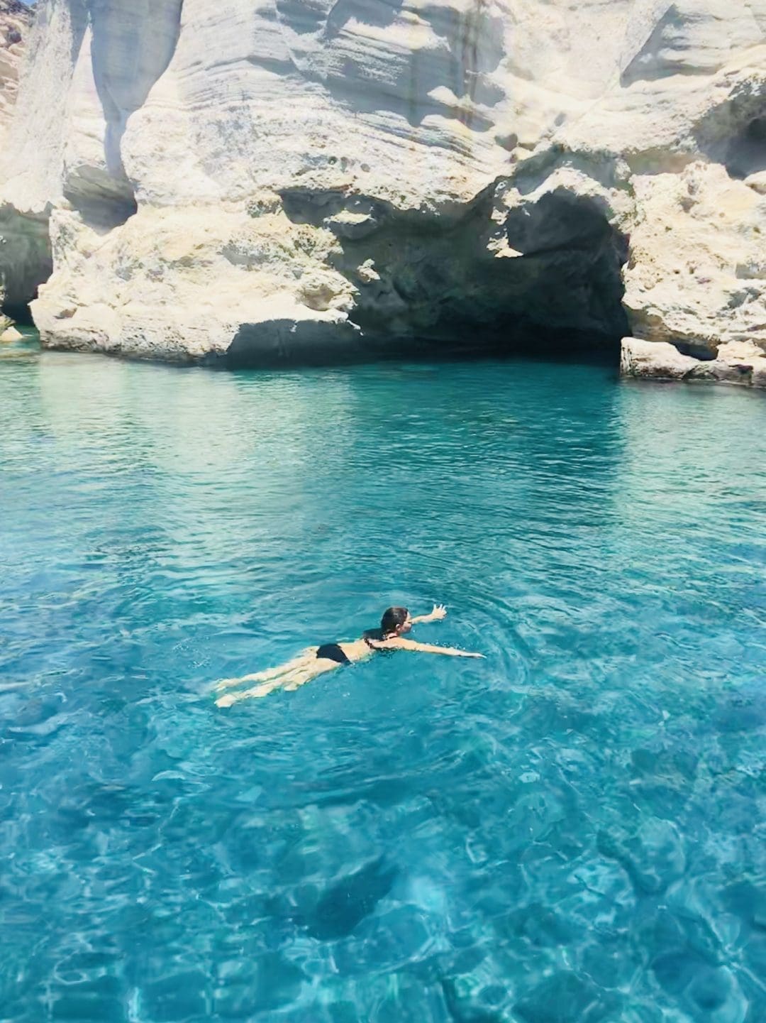 A person in a black swimsuit swims in clear, turquoise water near white rocky cliffs with a cave in the background, under bright sunlight.